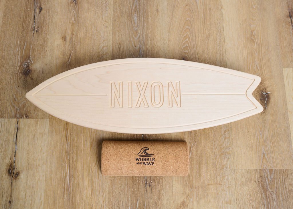 Wooden surfboard with 'Nixon' branding on a wooden surface, accompanied by a cork tube with 'Wobble and Wave' branding. Solid maple balance board featuring a bold capital name design. Supports balance, core strength, and adds a personal decor touch.