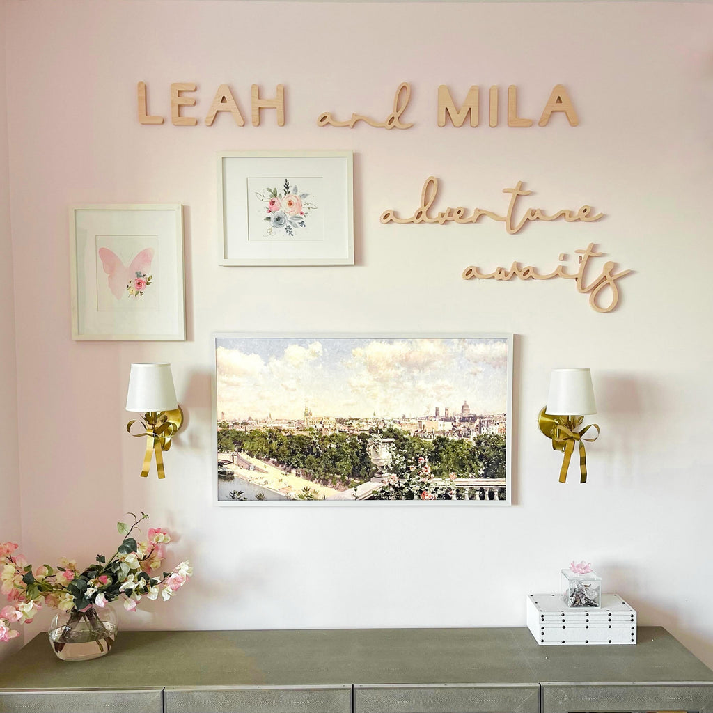 Wall decor with 'LEAH and MILA adventure awaits' on a pink wall with framed pictures and lamps.