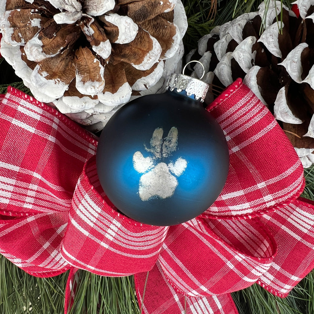 Decorative blue ornament with paw print on a red plaid bow, surrounded by pine cones and berries.