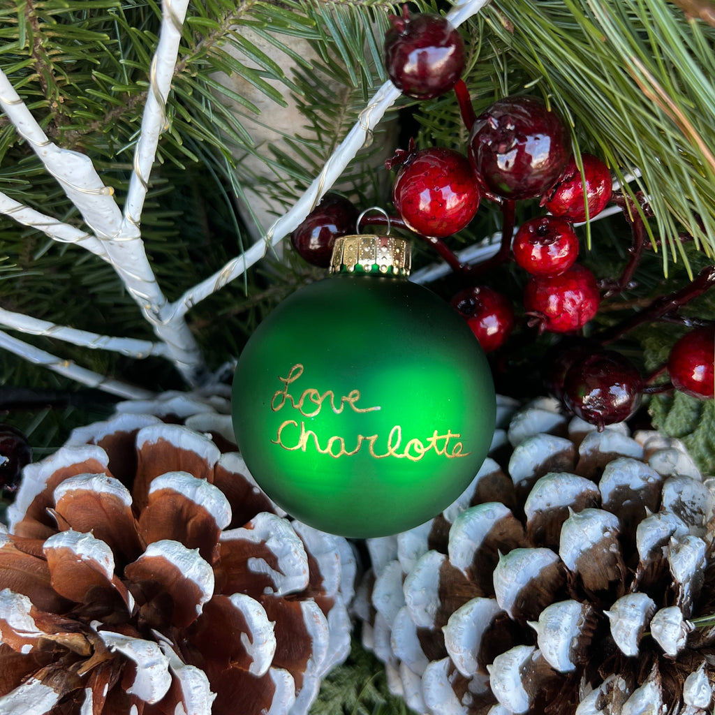 Decorative green ornament with 'Love Charlotte' text, surrounded by pinecones and berries on a Christmas tree.